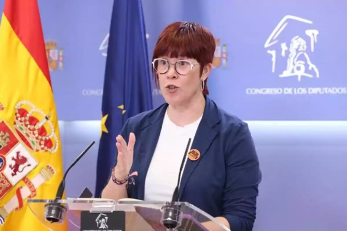Mujer con gafas y cabello corto rojizo hablando en una rueda de prensa en el Congreso de los Diputados de España, con banderas de España y la Unión Europea al fondo