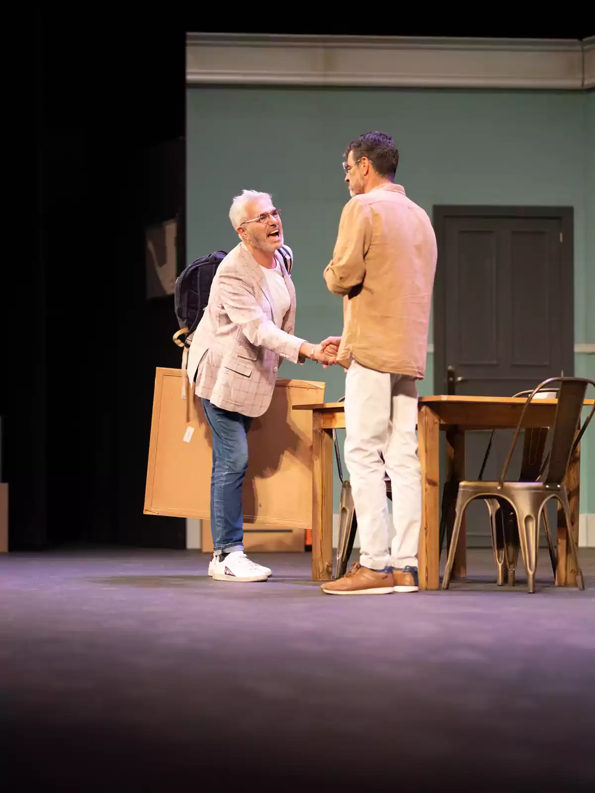 Dos hombres se saludan dándose la mano en un escenario de teatro junto a una mesa y sillas, uno de ellos lleva una mochila y un cuadro grande Dos hombres se saludan dándose la mano en un escenario de teatro junto a una mesa y sillas, uno de ellos lleva una mochila y un cuadro grande