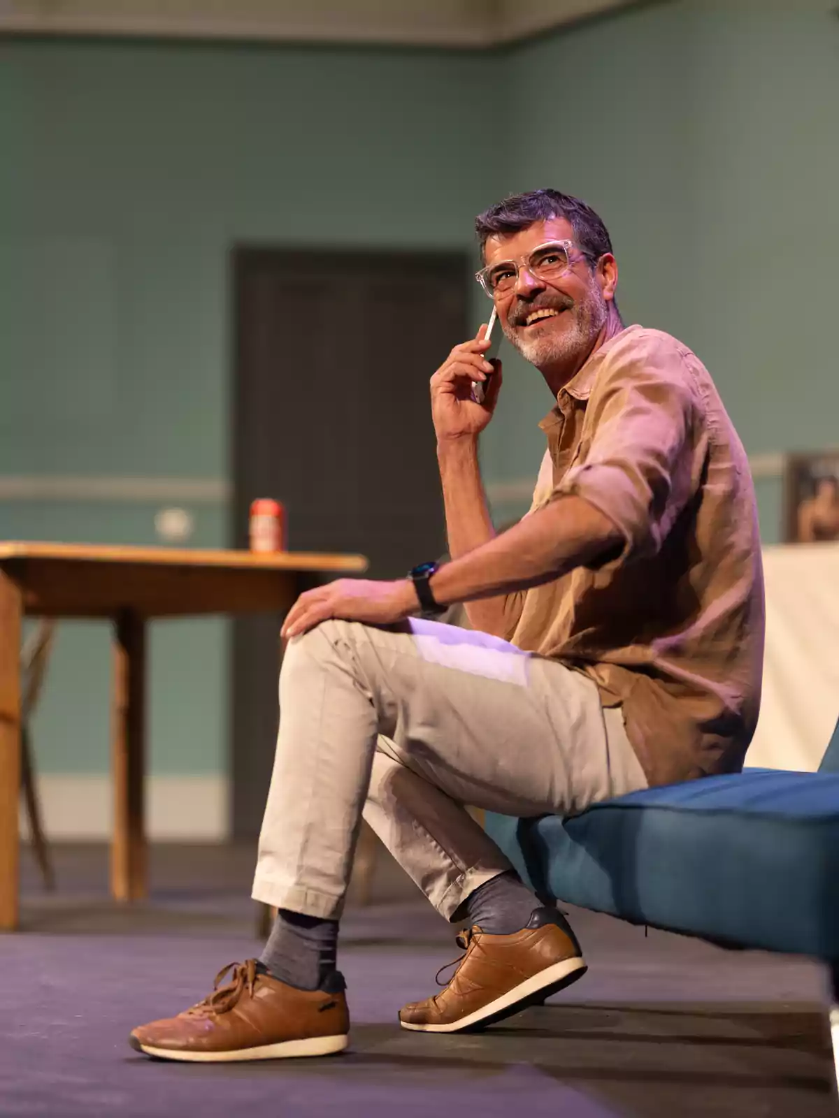 Hombre de mediana edad sentado en un sofá azul hablando por teléfono y sonriendo en un ambiente interior con una mesa de madera al fondo Hombre de mediana edad sentado en un sofá azul hablando por teléfono y sonriendo en un ambiente interior con una mesa de madera al fondo