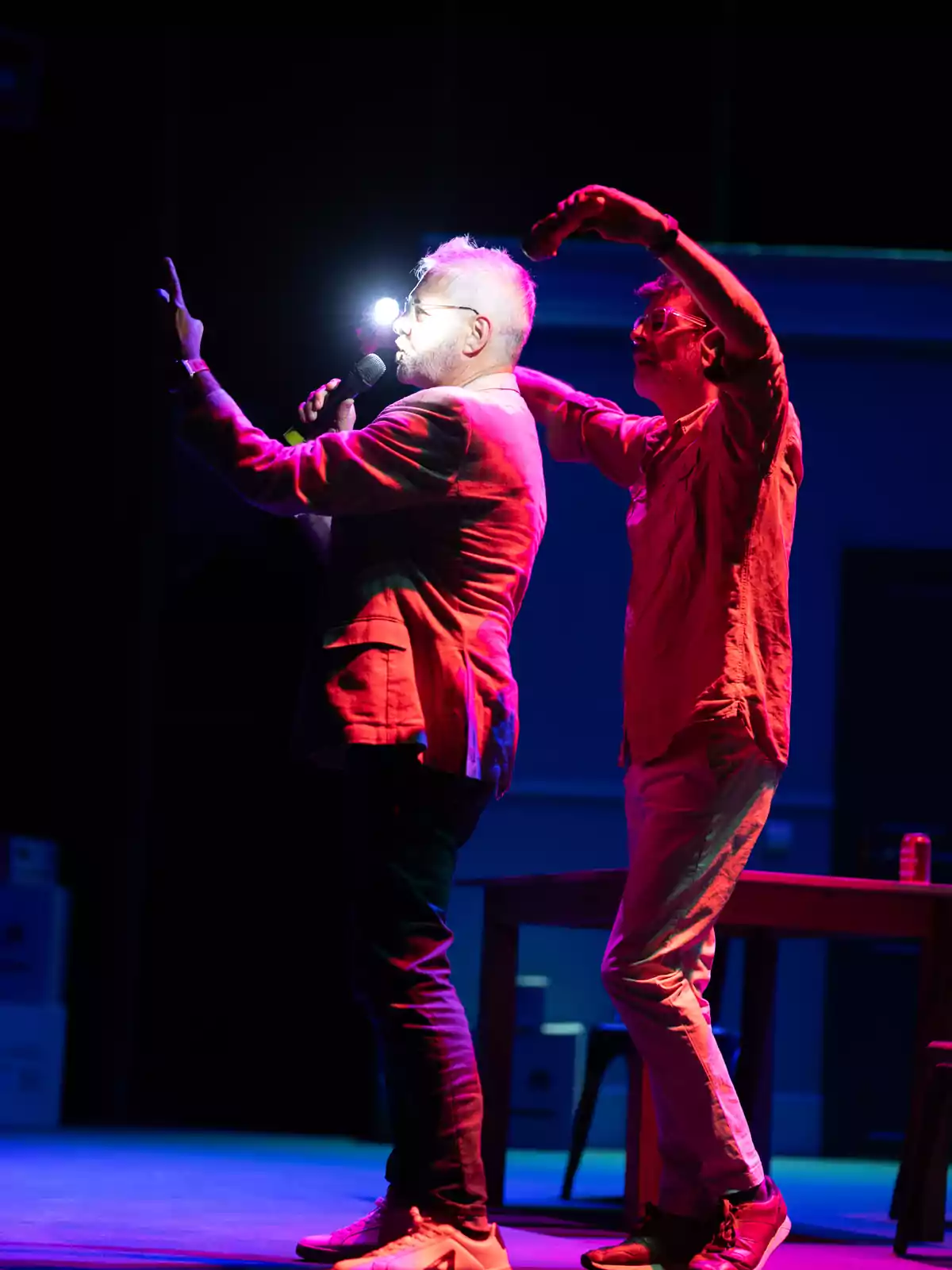 Dos hombres en el escenario iluminados con luces de colores, uno sostiene un micrófono y el otro está detrás de él con los brazos levantados Dos hombres en el escenario iluminados con luces de colores, uno sostiene un micrófono y el otro está detrás de él con los brazos levantados