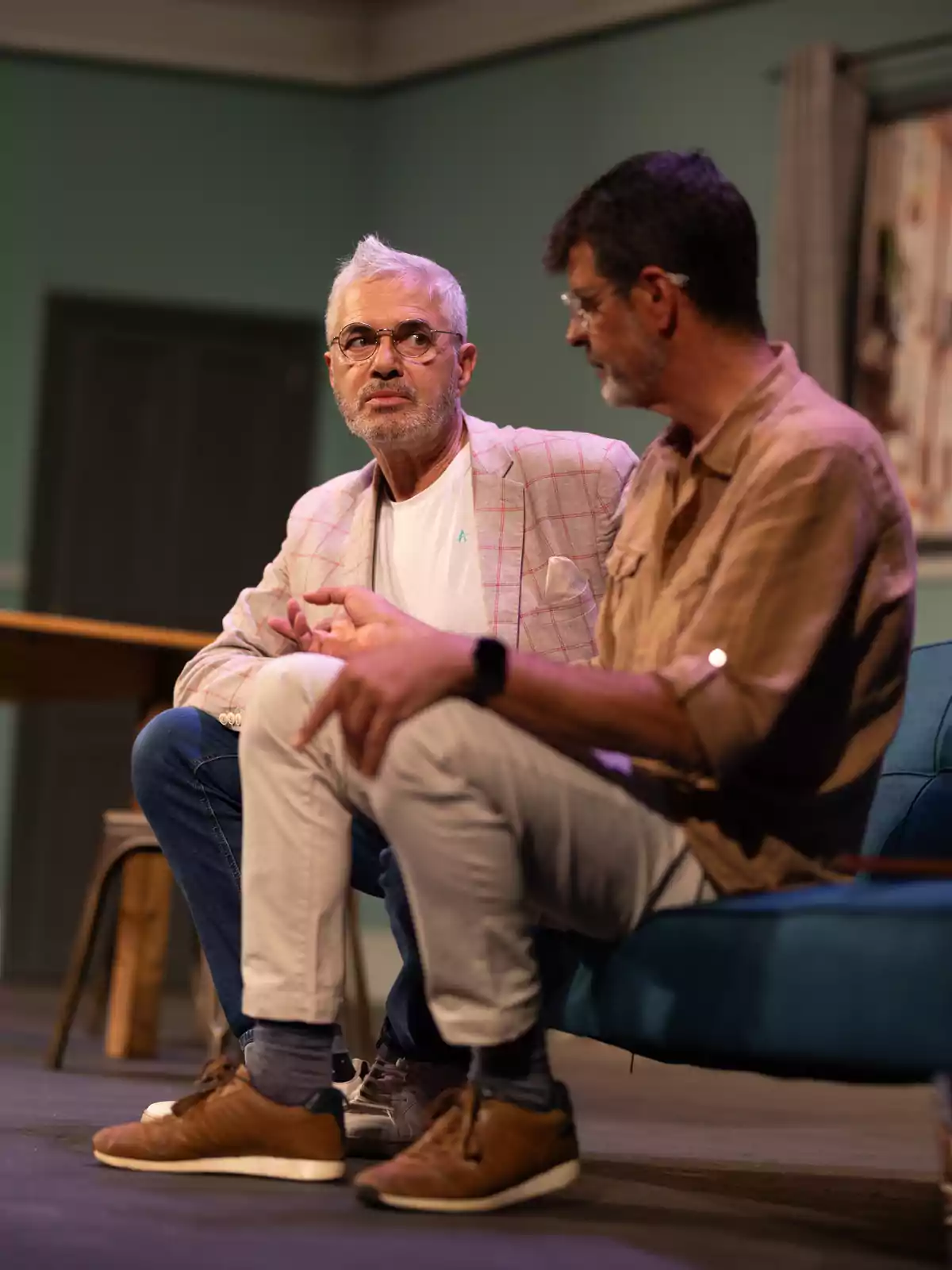 Dos hombres sentados en un sofá conversan en lo que parece ser una obra de teatro, uno de ellos lleva gafas y una chaqueta clara, el otro viste una camisa marrón y pantalones claros Dos hombres sentados en un sofá conversan en lo que parece ser una obra de teatro, uno de ellos lleva gafas y una chaqueta clara, el otro viste una camisa marrón y pantalones claros