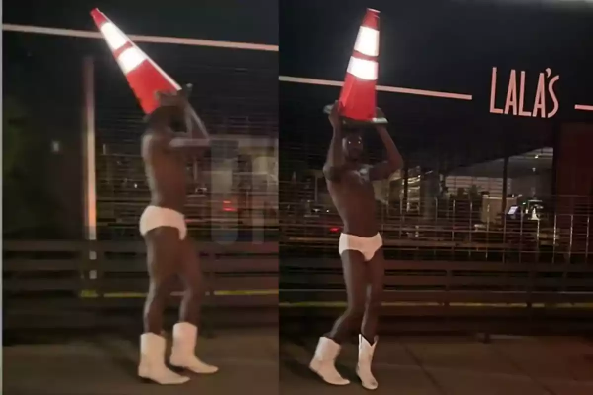 Persona caminando de noche en la calle usando solo ropa interior blanca, botas blancas y un cono de tráfico naranja en la cabeza frente a un local llamado Lala's Persona caminando de noche en la calle usando solo ropa interior blanca, botas blancas y un cono de tráfico naranja en la cabeza frente a un local llamado Lala's