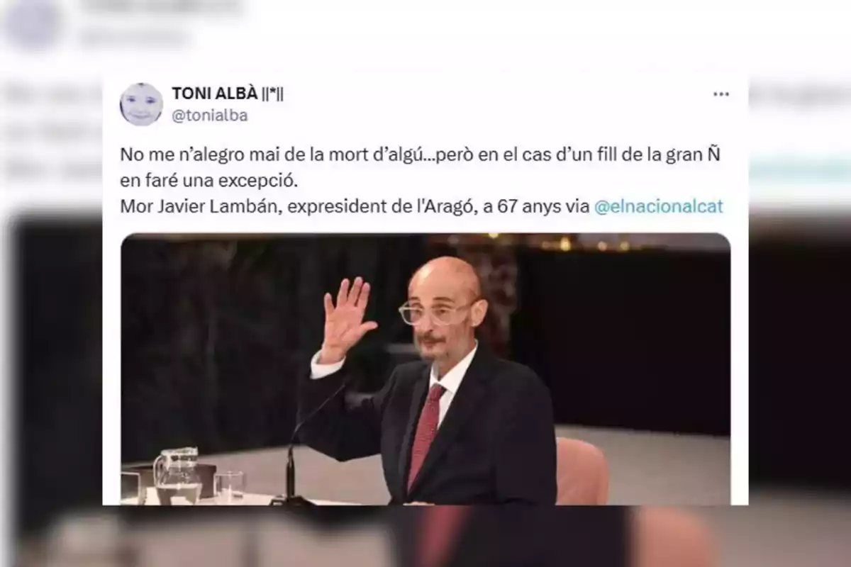 Captura de pantalla de un tuit en catalán con una foto de un hombre calvo con gafas y barba, vestido con traje y corbata, sentado en una mesa y levantando la mano Captura de pantalla de un tuit en catalán con una foto de un hombre calvo con gafas y barba, vestido con traje y corbata, sentado en una mesa y levantando la mano