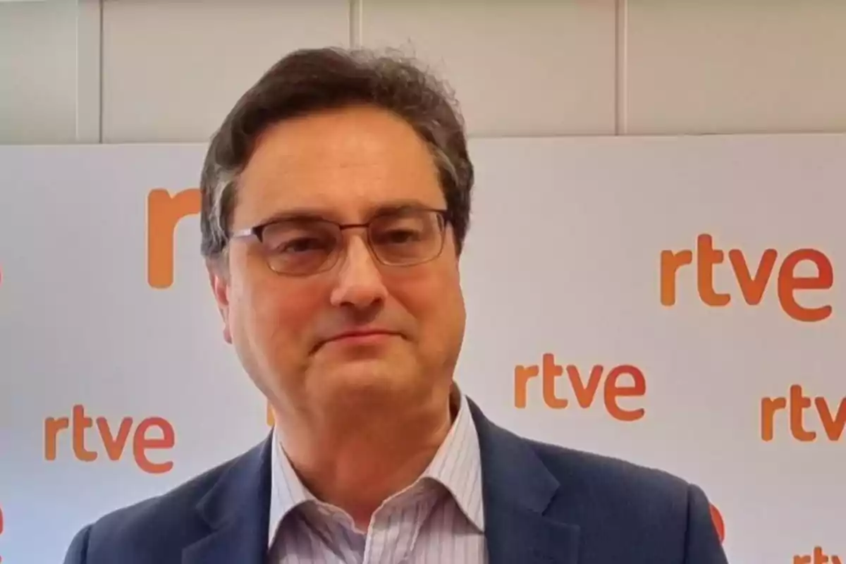 Hombre de cabello oscuro y gafas con saco azul posando frente a un fondo con el logo de rtve repetido