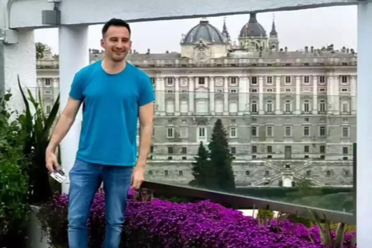 Hombre con camiseta azul posando en una terraza con flores moradas y un edificio histórico al fondo Hombre con camiseta azul posando en una terraza con flores moradas y un edificio histórico al fondo