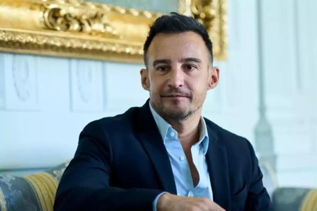 Hombre de cabello corto y barba ligera sentado en un sofá elegante con un cuadro dorado al fondo