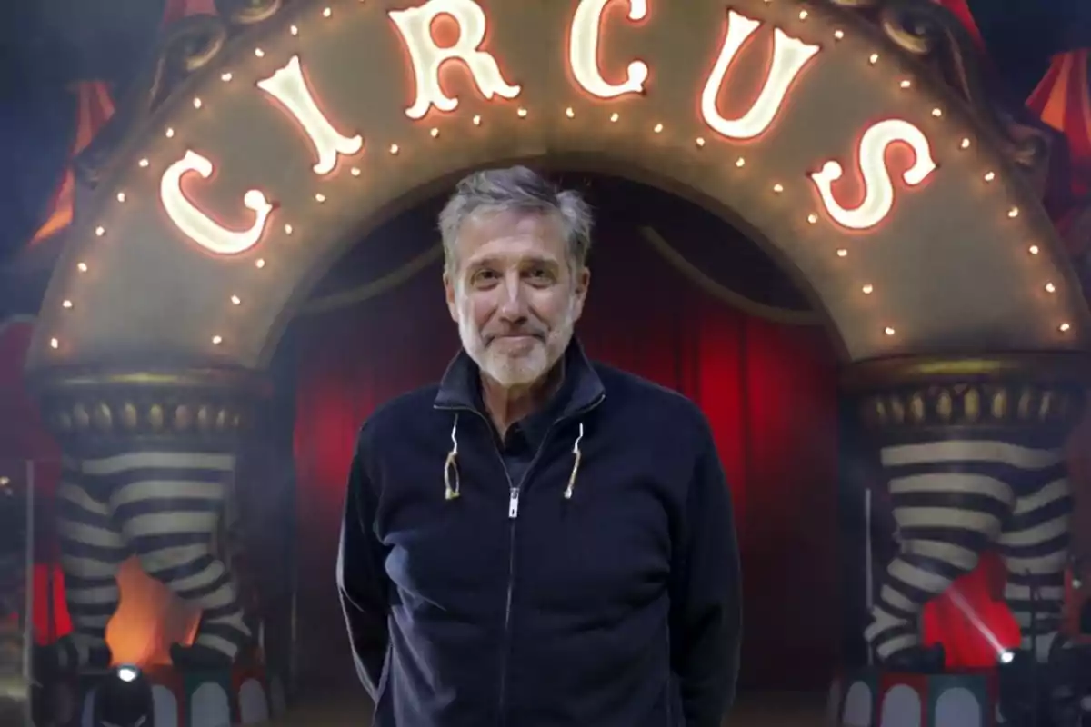 Hombre de cabello canoso y barba de pie frente a una entrada de circo iluminada con cortinas rojas de fondo