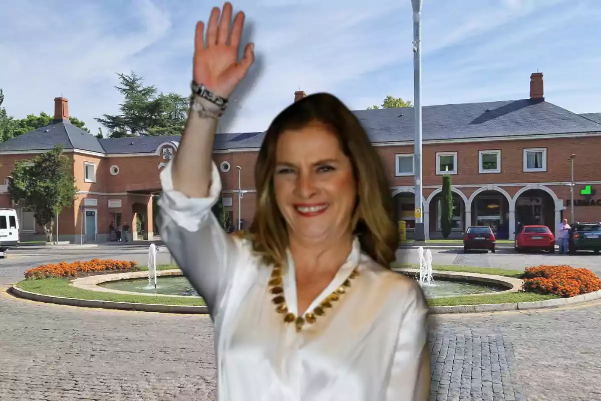 Mujer sonriente con camisa blanca y collar dorado levantando la mano frente a una plaza con fuente y edificio de ladrillo al fondo