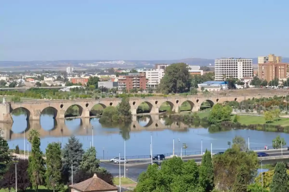 Puente romano de piedra sobre un río con edificios modernos al fondo y vegetación alrededor Puente romano de piedra sobre un río con edificios modernos al fondo y vegetación alrededor
