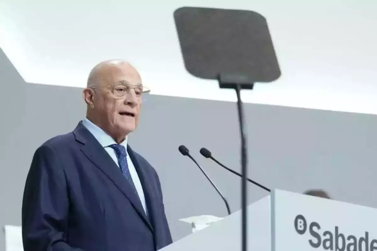Hombre mayor con gafas y traje azul hablando en un podio con el logo de Sabadell y un teleprompter frente a él