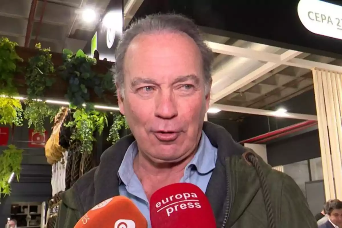 Hombre de mediana edad hablando frente a varios micrófonos en un evento interior con plantas decorativas al fondo Hombre de mediana edad hablando frente a varios micrófonos en un evento interior con plantas decorativas al fondo
