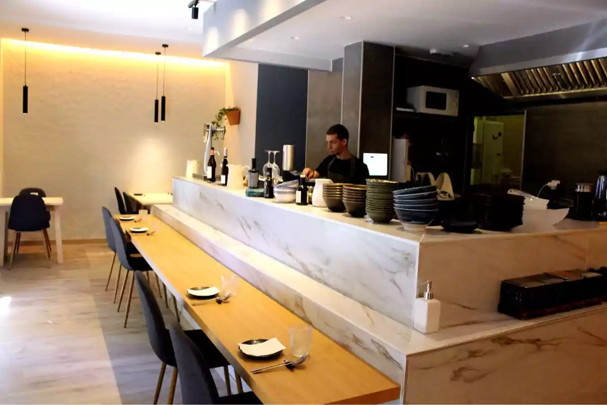 Restaurante moderno con barra de mármol, mesas de madera, sillas oscuras y un chef trabajando detrás del mostrador