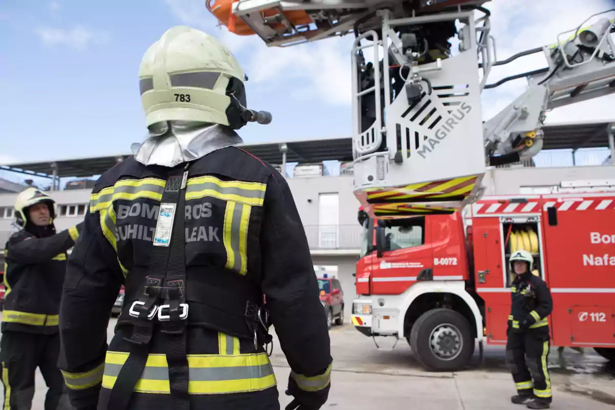 Varios bomberos con uniforme y casco trabajan junto a un camión de bomberos rojo equipado con una escalera mecánica en una zona urbana Varios bomberos con uniforme y casco trabajan junto a un camión de bomberos rojo equipado con una escalera mecánica en una zona urbana