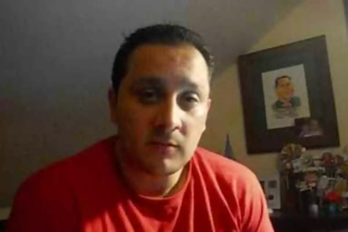 Hombre con camiseta roja sentado en una habitación con pared inclinada y un retrato enmarcado al fondo Hombre con camiseta roja sentado en una habitación con pared inclinada y un retrato enmarcado al fondo