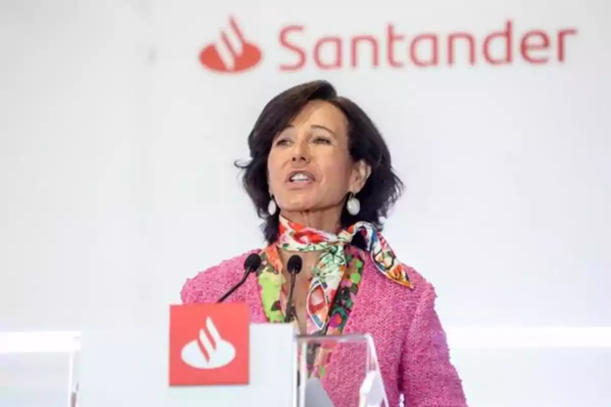 Mujer con chaqueta rosa y pañuelo colorido hablando en un podio con el logo de Santander al fondo