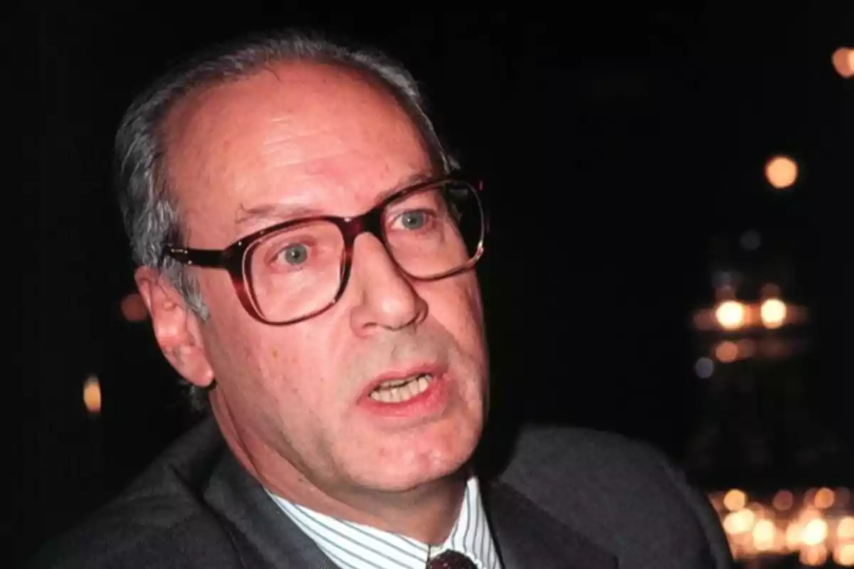 Hombre mayor con gafas grandes y cabello canoso vestido con traje oscuro y corbata en un ambiente oscuro con luces desenfocadas al fondo Hombre mayor con gafas grandes y cabello canoso vestido con traje oscuro y corbata en un ambiente oscuro con luces desenfocadas al fondo