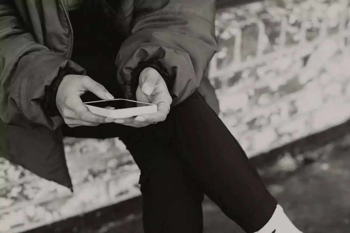 Persona sentada sosteniendo un teléfono móvil con ambas manos, usando chaqueta y pantalones oscuros, imagen en blanco y negro. Persona sentada sosteniendo un teléfono móvil con ambas manos, usando chaqueta y pantalones oscuros, imagen en blanco y negro.
