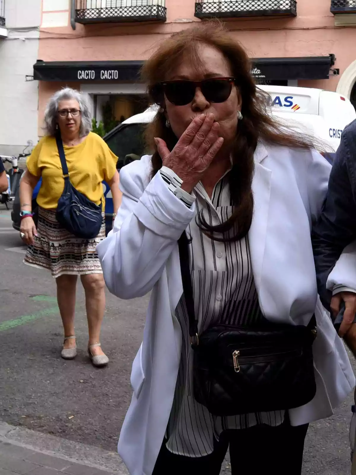 Mujer con gafas de sol y chaqueta blanca lanzando un beso mientras camina por la calle, con otra mujer al fondo y un edificio detrás