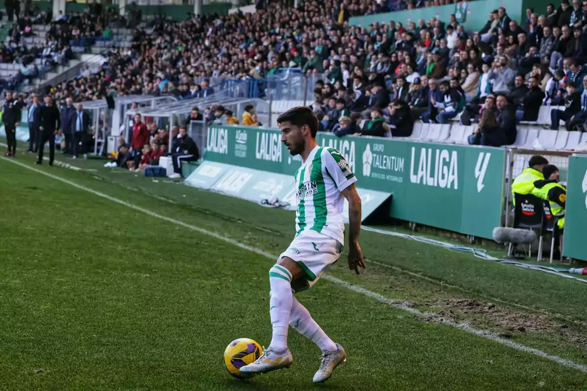 Futbolista con uniforme blanco y verde controlando el balón cerca de la línea de banda en un estadio lleno de espectadores Futbolista con uniforme blanco y verde controlando el balón cerca de la línea de banda en un estadio lleno de espectadores