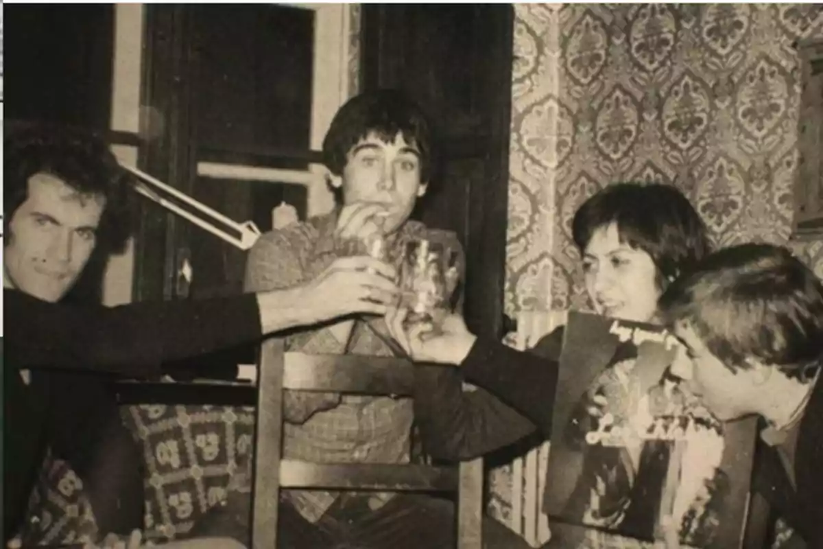 Cuatro personas jóvenes brindan con vasos en una habitación con papel tapiz decorativo y una de ellas sostiene un disco de vinilo Cuatro personas jóvenes brindan con vasos en una habitación con papel tapiz decorativo y una de ellas sostiene un disco de vinilo