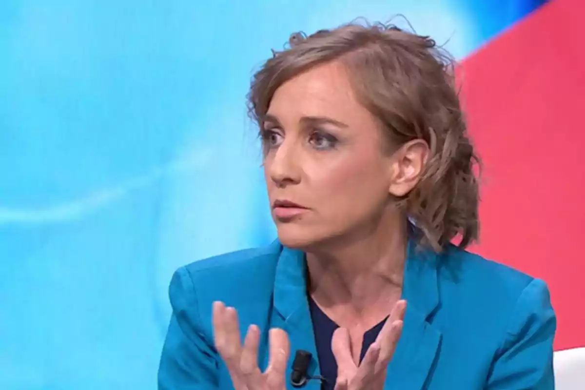 Mujer de cabello corto y rubio vestida con saco azul hablando y gesticulando en un estudio de televisión con fondo azul y rojo Mujer de cabello corto y rubio vestida con saco azul hablando y gesticulando en un estudio de televisión con fondo azul y rojo