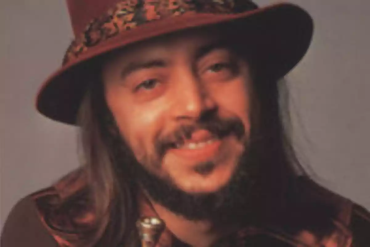 Hombre con barba y bigote usando sombrero marrón y camisa oscura mientras sonríe a la cámara