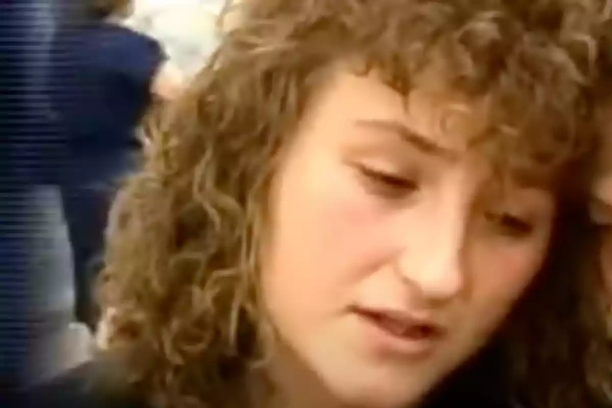 Mujer joven de cabello rizado y expresión seria mirando hacia abajo en un entorno con personas al fondo Mujer joven de cabello rizado y expresión seria mirando hacia abajo en un entorno con personas al fondo