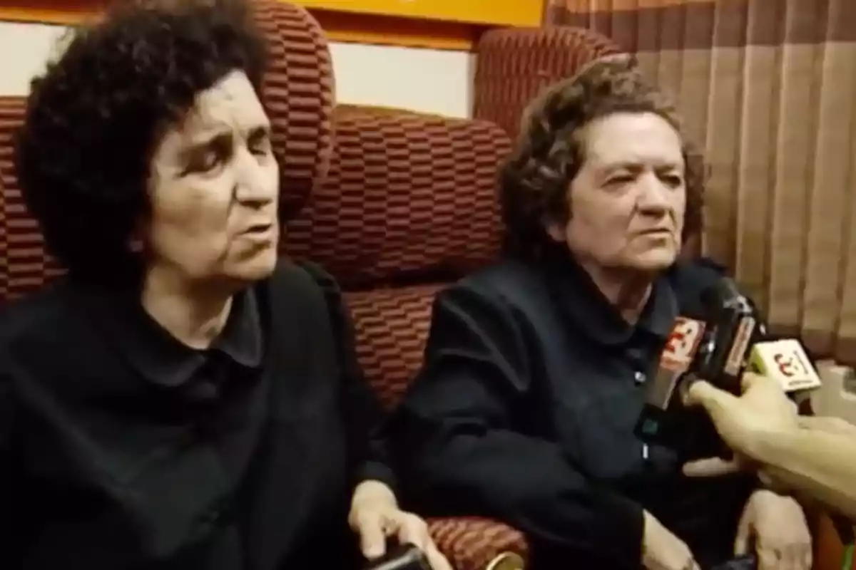 Dos mujeres mayores sentadas en sillones rojos mientras son entrevistadas por reporteros que sostienen micrófonos frente a ellas Dos mujeres mayores sentadas en sillones rojos mientras son entrevistadas por reporteros que sostienen micrófonos frente a ellas
