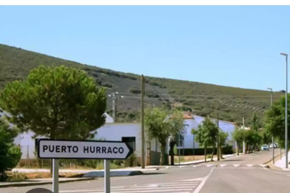 Señal de tráfico que indica la dirección hacia Puerto Hurraco en una carretera rodeada de árboles y colinas bajo un cielo despejado Señal de tráfico que indica la dirección hacia Puerto Hurraco en una carretera rodeada de árboles y colinas bajo un cielo despejado
