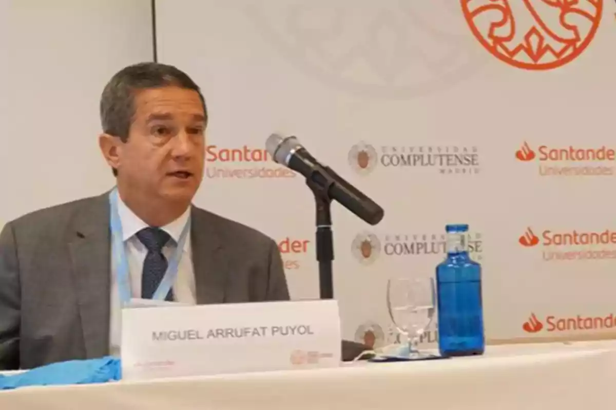 Hombre de traje hablando en una conferencia con un micrófono frente a él y una botella de agua sobre la mesa Hombre de traje hablando en una conferencia con un micrófono frente a él y una botella de agua sobre la mesa