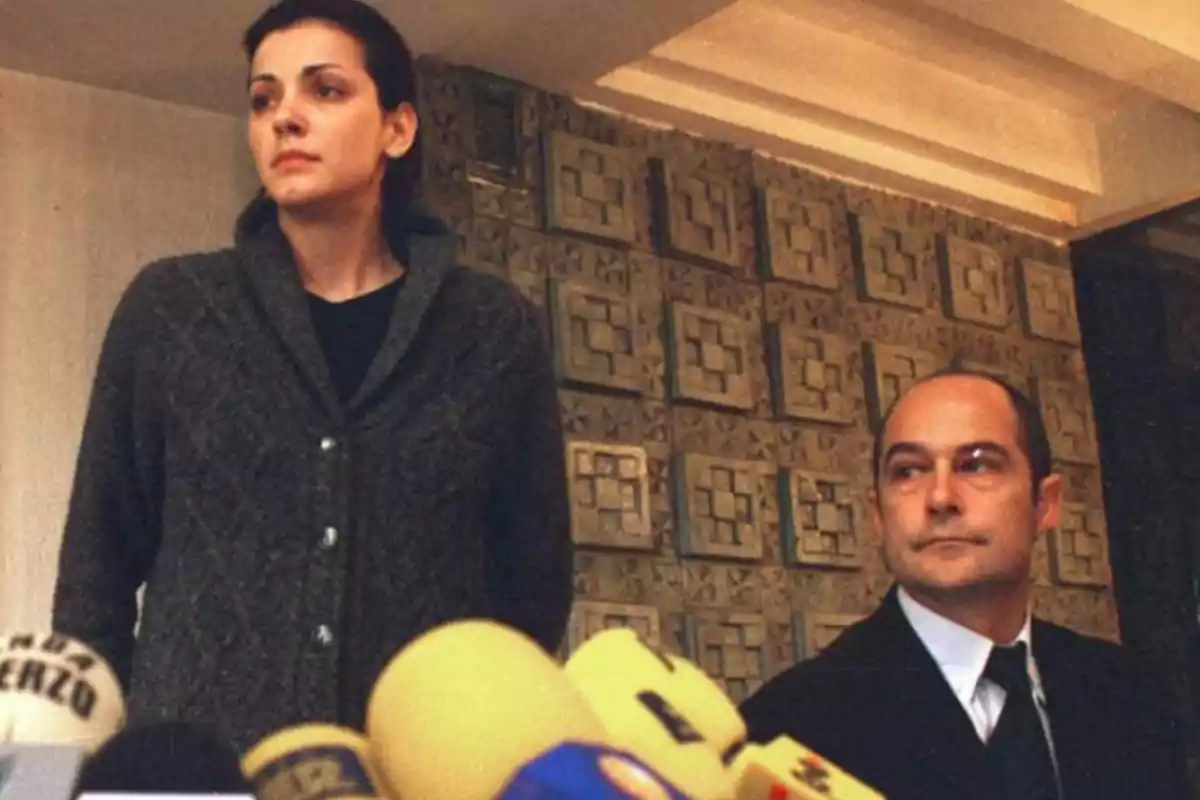 Una mujer de pie y un hombre sentado frente a varios micrófonos en una conferencia de prensa en un interior con pared decorativa de fondo Una mujer de pie y un hombre sentado frente a varios micrófonos en una conferencia de prensa en un interior con pared decorativa de fondo