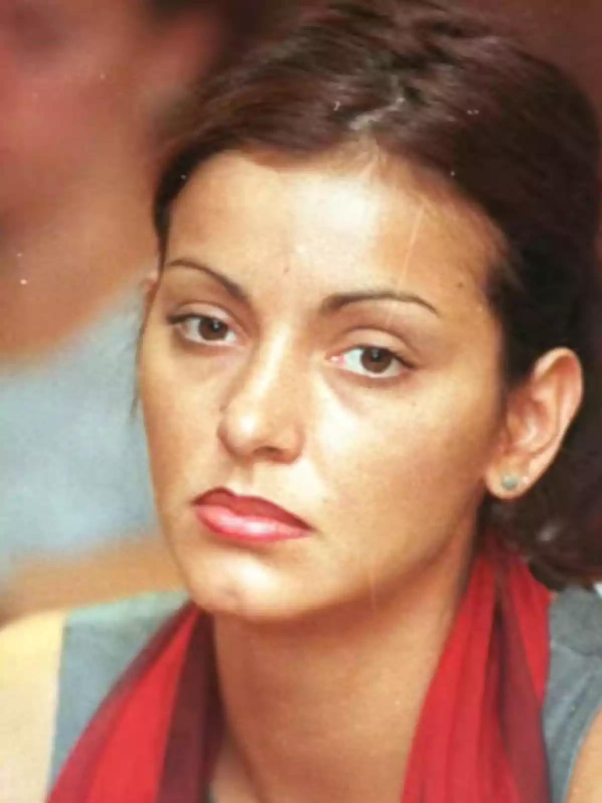 Mujer de cabello oscuro con expresión seria, usando una bufanda roja y aretes pequeños Mujer de cabello oscuro con expresión seria, usando una bufanda roja y aretes pequeños