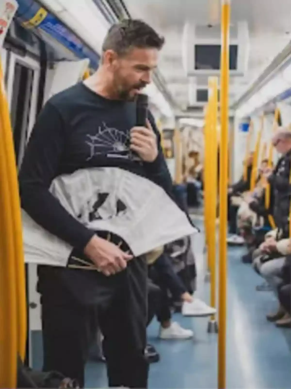 Hombre hablando por micrófono y sosteniendo un abanico grande dentro de un vagón de metro mientras los pasajeros observan Hombre hablando por micrófono y sosteniendo un abanico grande dentro de un vagón de metro mientras los pasajeros observan