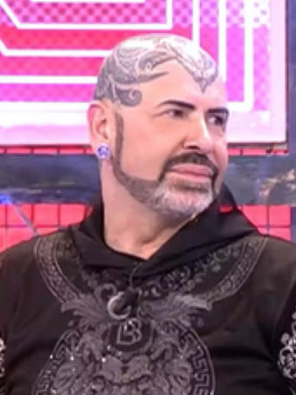 Hombre calvo con tatuajes en la cabeza, barba perfilada y ropa oscura con detalles decorativos, sentado en un set de televisión con fondo rosa y rojo. Hombre calvo con tatuajes en la cabeza, barba perfilada y ropa oscura con detalles decorativos, sentado en un set de televisión con fondo rosa y rojo.