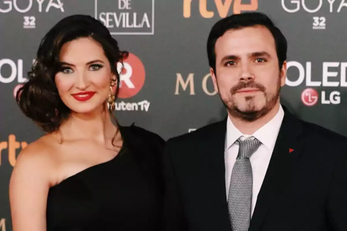 Una pareja elegantemente vestida posa en una alfombra roja frente a un fondo con logotipos de patrocinadores y eventos.