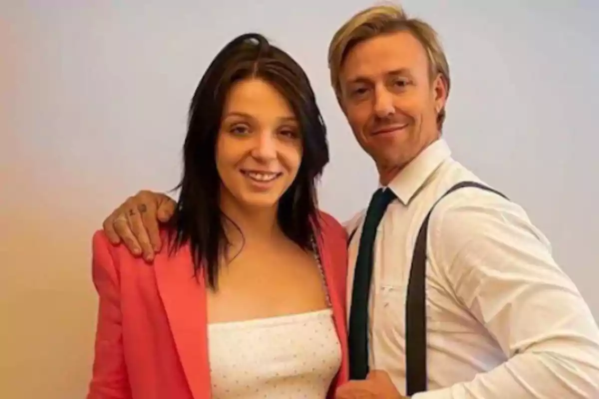 Una mujer con chaqueta rosa y un hombre con camisa blanca y tirantes posan juntos sonriendo frente a una pared clara Una mujer con chaqueta rosa y un hombre con camisa blanca y tirantes posan juntos sonriendo frente a una pared clara