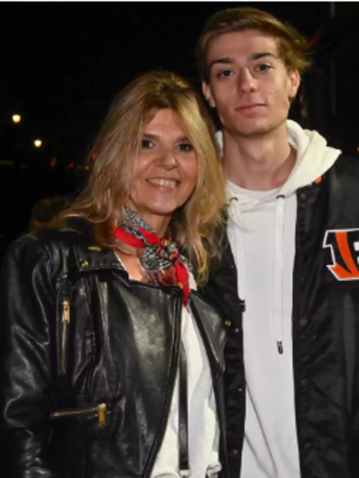Dos personas posan juntas y sonríen, una mujer con chaqueta de cuero negra y un joven con chaqueta deportiva negra y sudadera blanca, ambos en un ambiente nocturno. Dos personas posan juntas y sonríen, una mujer con chaqueta de cuero negra y un joven con chaqueta deportiva negra y sudadera blanca, ambos en un ambiente nocturno.