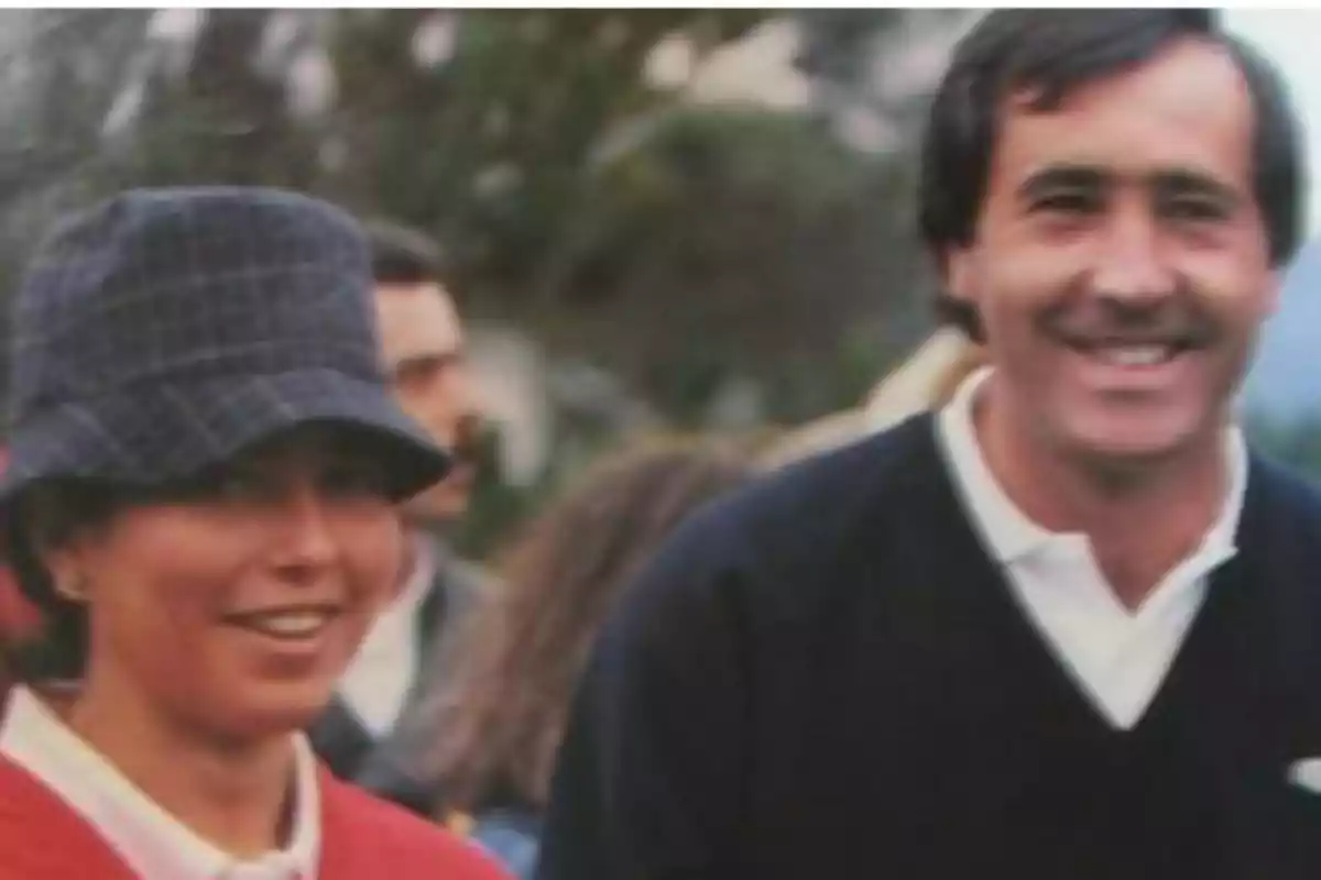 Dos personas sonrientes al aire libre, una lleva un sombrero de cuadros y la otra viste un suéter oscuro sobre una camisa blanca Dos personas sonrientes al aire libre, una lleva un sombrero de cuadros y la otra viste un suéter oscuro sobre una camisa blanca