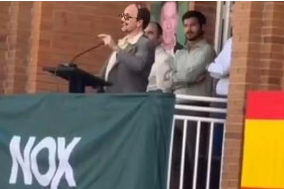 Un hombre con gafas habla en un podio mientras otras personas lo observan y una lona verde con la palabra NOX cuelga al frente. Un hombre con gafas habla en un podio mientras otras personas lo observan y una lona verde con la palabra NOX cuelga al frente.