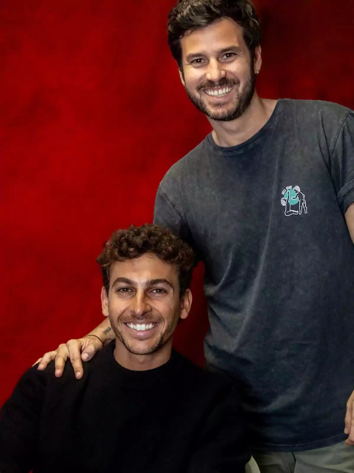 Dos hombres sonríen frente a un fondo rojo, uno está sentado y el otro de pie con la mano sobre su hombro Dos hombres sonríen frente a un fondo rojo, uno está sentado y el otro de pie con la mano sobre su hombro
