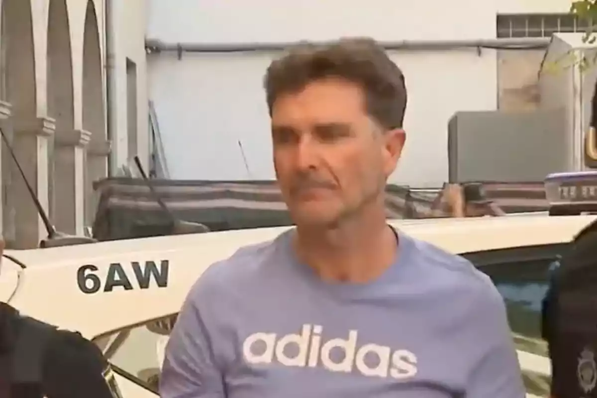 Hombre de cabello corto y canoso con camiseta lila de la marca Adidas siendo escoltado por la policía frente a un vehículo patrulla Hombre de cabello corto y canoso con camiseta lila de la marca Adidas siendo escoltado por la policía frente a un vehículo patrulla
