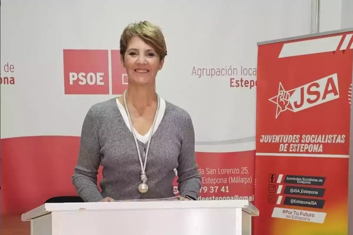Mujer sonriente de pie detrás de un atril en un evento de Juventudes Socialistas de Estepona con carteles de PSOE y JSA al fondo Mujer sonriente de pie detrás de un atril en un evento de Juventudes Socialistas de Estepona con carteles de PSOE y JSA al fondo