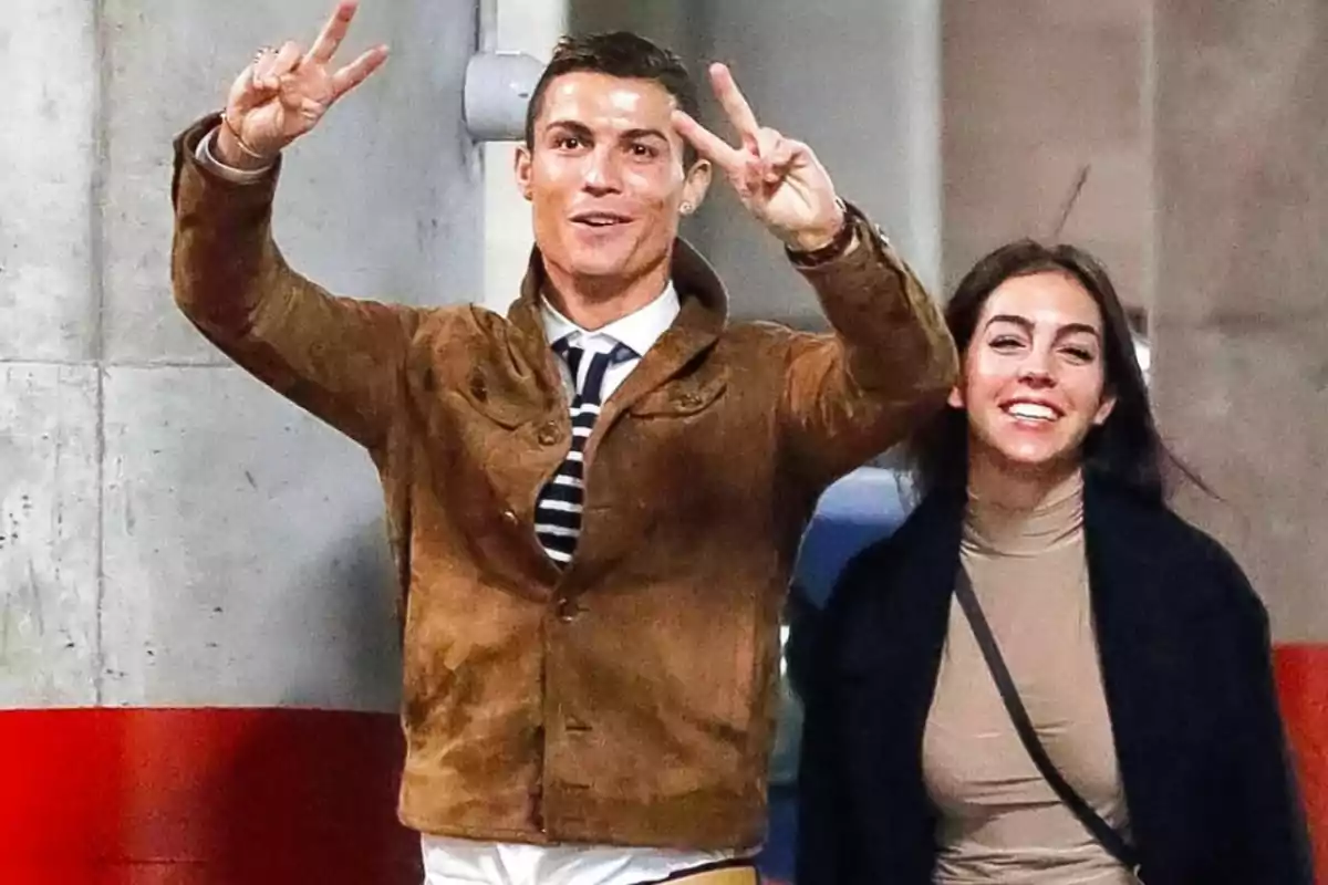 Un hombre sonriente hace el signo de la paz con ambas manos mientras camina junto a una mujer que también sonríe en un estacionamiento.