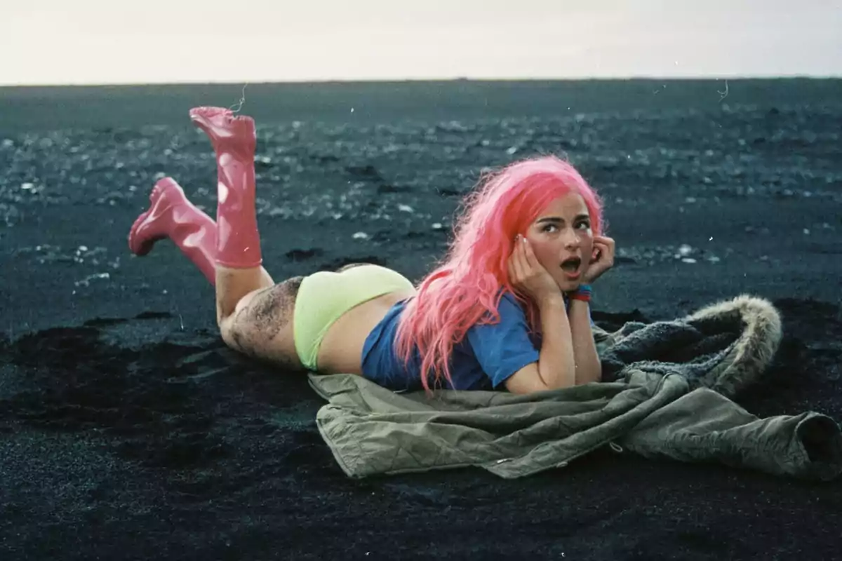 Persona con cabello rosa, camiseta azul, botas rosas y ropa interior verde acostada sobre una chaqueta en la arena oscura de una playa