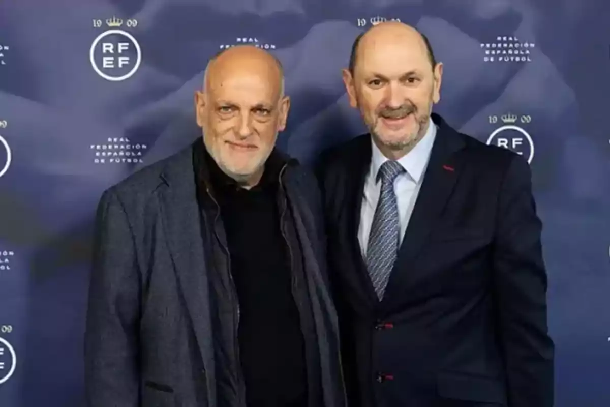 Dos hombres posan juntos frente a un fondo azul con logotipos de la Real Federación Española de Fútbol