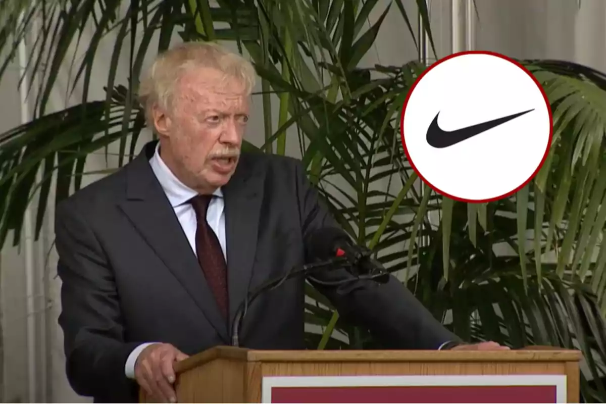 Hombre de cabello canoso y bigote hablando en un podio con plantas de fondo y el logotipo de Nike en la esquina superior derecha