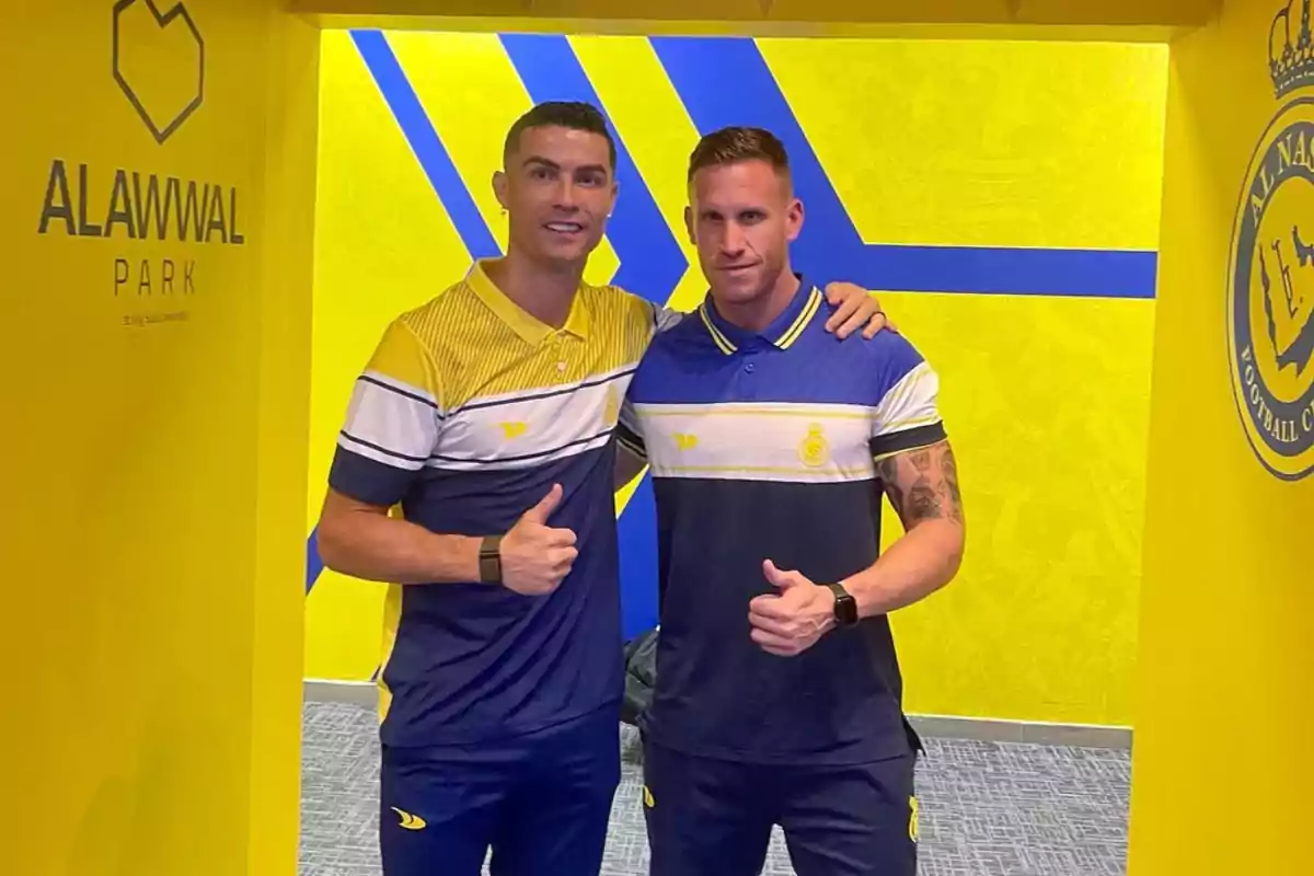 Dos hombres con uniforme deportivo posan juntos en un pasillo amarillo con logotipos y fondo de Al Nassr, ambos sonríen y levantan el pulgar.