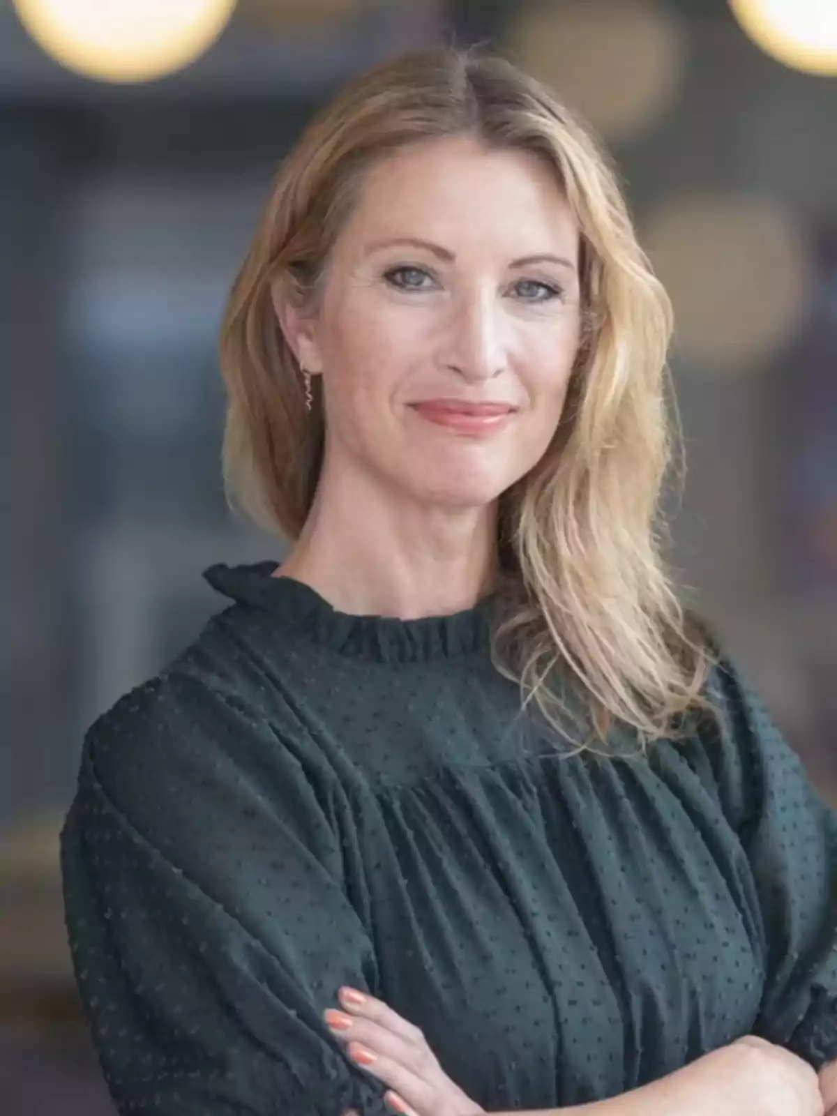 Mujer rubia de mediana edad con cabello suelto y ondulado, vestida con blusa oscura de manga larga, posando con los brazos cruzados y una expresión confiada Mujer rubia de mediana edad con cabello suelto y ondulado, vestida con blusa oscura de manga larga, posando con los brazos cruzados y una expresión confiada