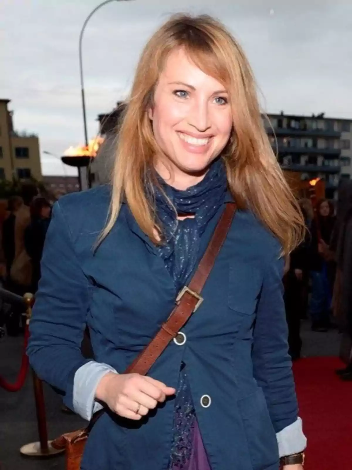 Mujer rubia sonriente con chaqueta azul y bufanda caminando por una alfombra roja al aire libre Mujer rubia sonriente con chaqueta azul y bufanda caminando por una alfombra roja al aire libre