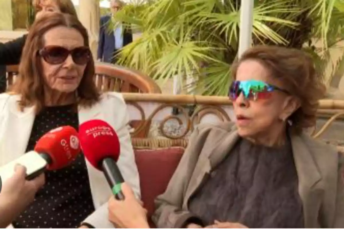 Dos mujeres mayores sentadas en un sofá al aire libre, una lleva gafas de sol oscuras y la otra gafas de sol llamativas de colores, ambas están siendo entrevistadas por periodistas que sostienen micrófonos rojos Dos mujeres mayores sentadas en un sofá al aire libre, una lleva gafas de sol oscuras y la otra gafas de sol llamativas de colores, ambas están siendo entrevistadas por periodistas que sostienen micrófonos rojos